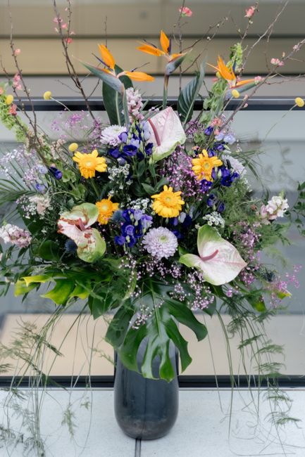 A vibrant floral arrangement in a black glass vase featuring a mix of fresh flowers including large white calla lilies with pink streaks, bright yellow gerbera daisies, deep blue delphiniums, small wh