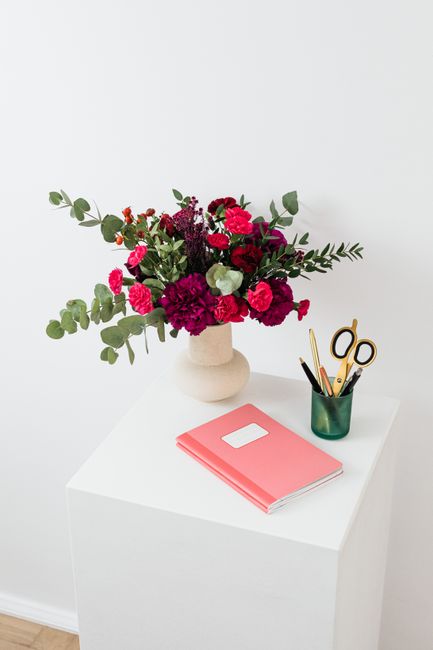 A vibrant floral arrangement featuring deep pink carnations, purple and red spray roses, and white green-flowered blooms, accented with eucalyptus leaves, all placed in a simple, cream-colored ceramic