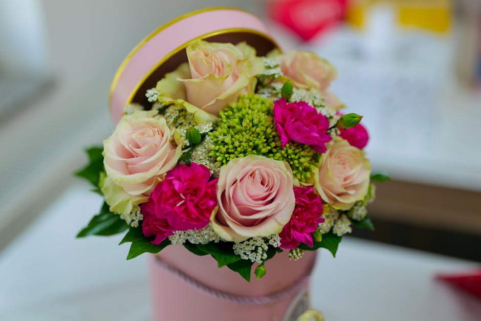 A round floral arrangement in a pink hatbox, featuring pale pink roses, vibrant pink carnations, small white and green filler flowers, and lush green leaves, arranged in a compact, symmetrical style. 