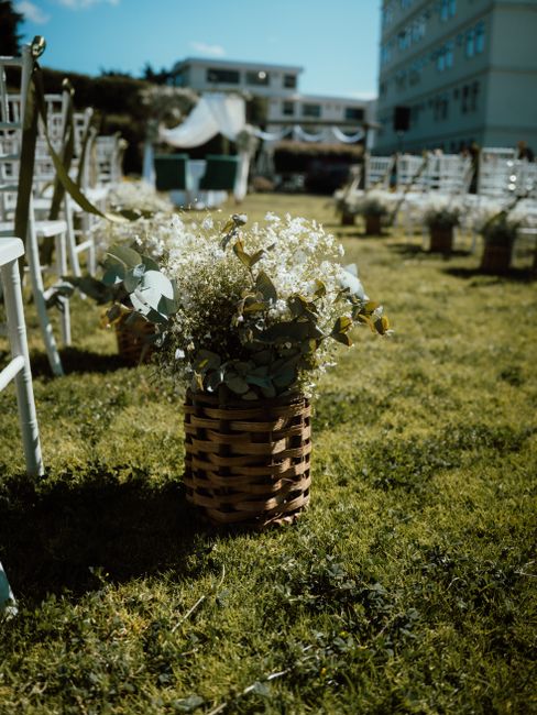 A floral arrangement featuring predominantly white flowers, including delicate baby's breath and soft eucalyptus leaves, arranged in a woven wicker basket. The bouquet is placed on a grassy area at Ea