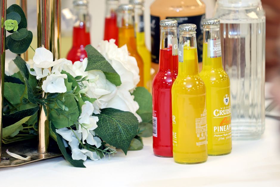 A floral arrangement featuring white hydrangeas with lush green leaves, placed next to a collection of colorful bottled beverages including red and yellow drinks, on a white surface. The hydrangeas di