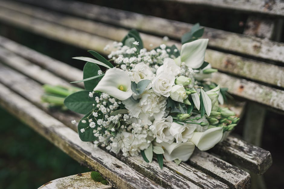 A bouquet of fresh white flowers rests on a weathered wooden bench, featuring elegant calla lilies, roses, and spray carnations, accented with eucalyptus and baby's breath. The flowers are arranged in