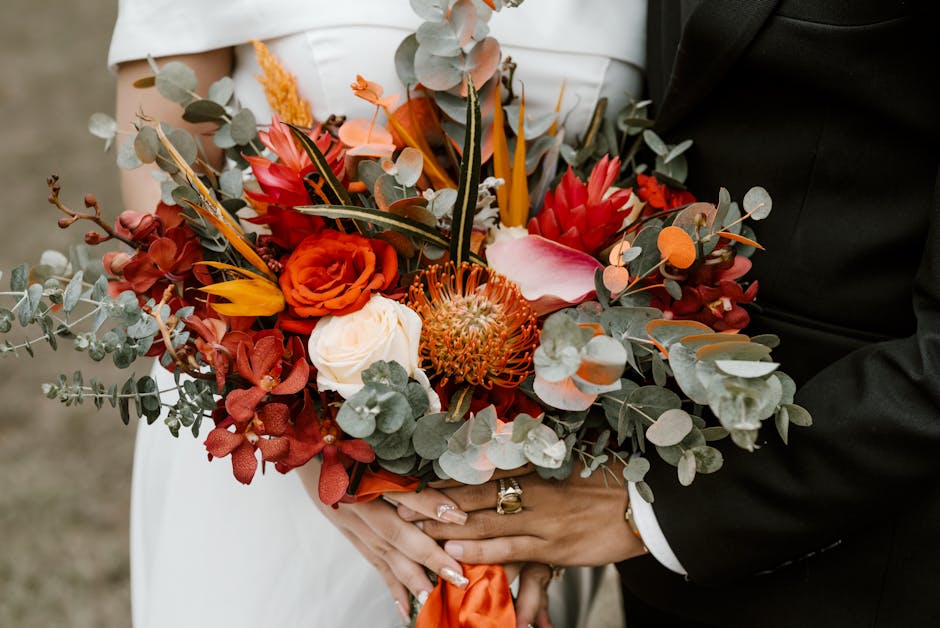 A detailed floral bouquet held by a bride and groom, featuring a mix of vibrant colours including deep red roses, orange proteas, blush pink calla lilies, and white roses, complemented by muted green