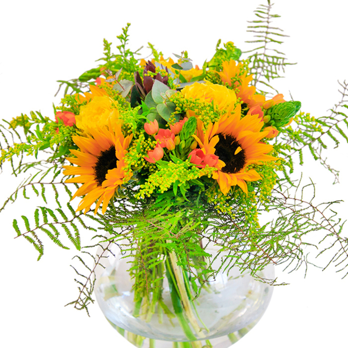 At first glance, this bouquet radiates the bright optimism of a sunlit afternoon in Dagenham, where sunflowers and garden roses come alive in harmonious celebration. Nestled within a clear, round vase, the blooms trace arcs of yellow and gold that glow softly against a snowy background-suggesting the open skies above the borough during a clear late-summer day. Gorgeously spiraled yellow roses complement the star-like sunflowers, while glossy red hypericum berries glimmer within the foliage, creating flashes of colour that recall the bustling atmosphere at Dagenham Market. Wisps of ferns and cascading greenery bring natural movement, softening the edges and adding a sense of wild abundance seen in community parks. Eucalyptus sprigs, tinged with bluish-green, weave gently through goldenrod clusters and delicate filler, their subtle fragrance melding with the faint sweetness of the roses. Submerged stems, crisply visible in pure water, speak to both freshness and transparency-values cherished by local residents. This bouquet, crafted by Florist Dagenham for milestones and heartfelt occasions, is a sensory tribute: to joy, friendship, and the enduring beauty of the area's green spaces. Each petal, leaf, and berry is placed with intention, creating not just a floral gift, but an experience that connects nature's artistry with the warmth of home.